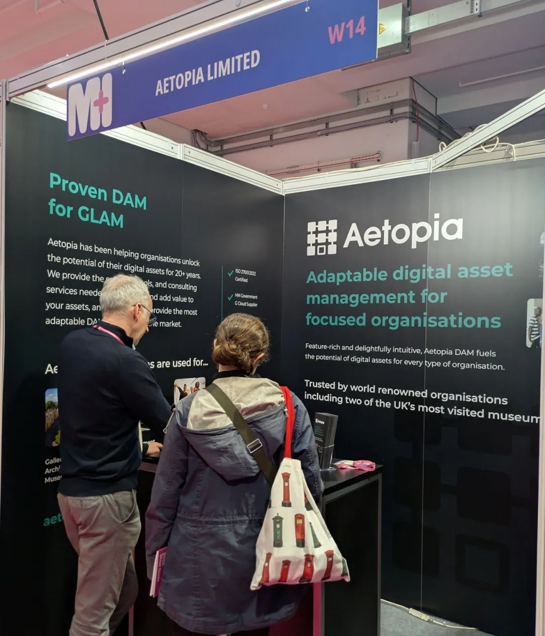Aetopia's Stand W14 at the Museums + Heritage Show, with a team member and a visitor in conversation beneath 'Proven DAM for GLAM' signage